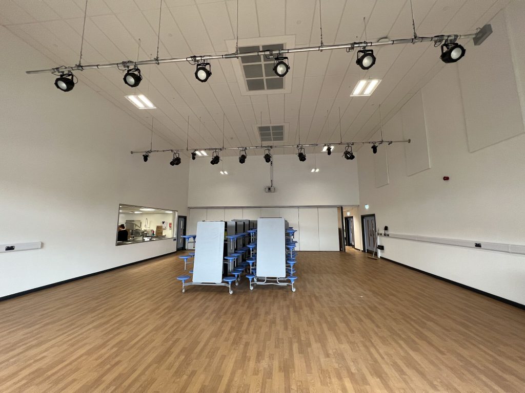 A school hall with fold away tables stacked in the middle of the room and two sets of lighting rigs suspended from the ceiling.