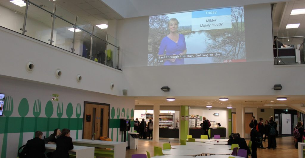 Image of school canteen with a small number of students sitting at tables while others line up for food