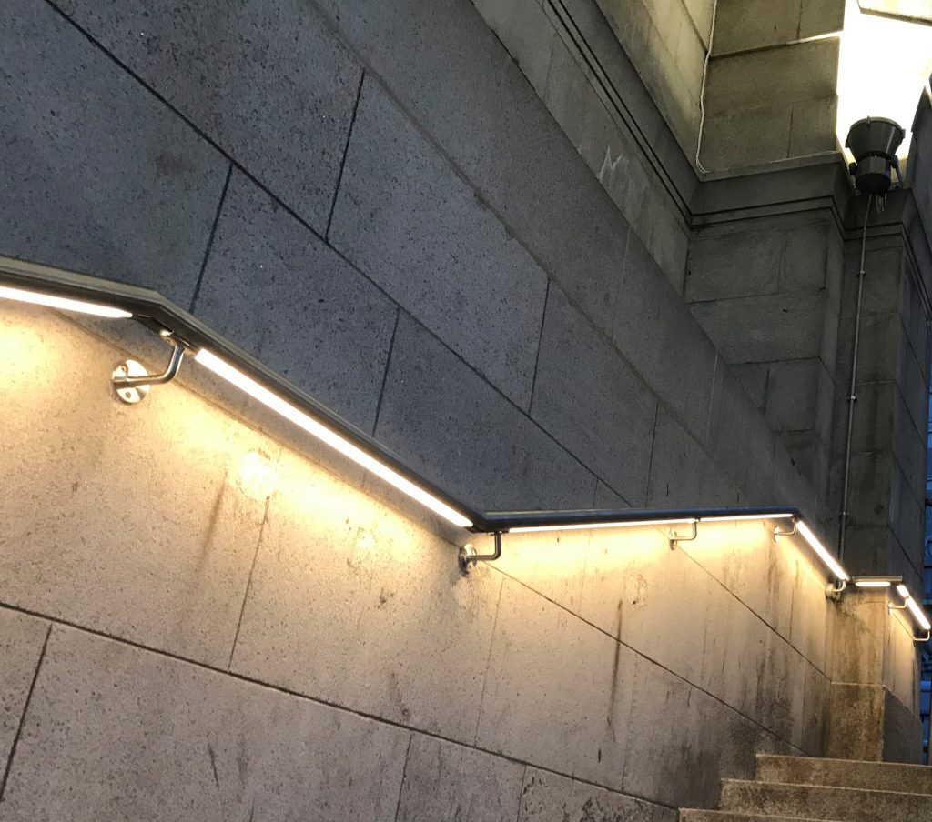 Photo of an illuminated handrail going up alongside stairs at a train station.