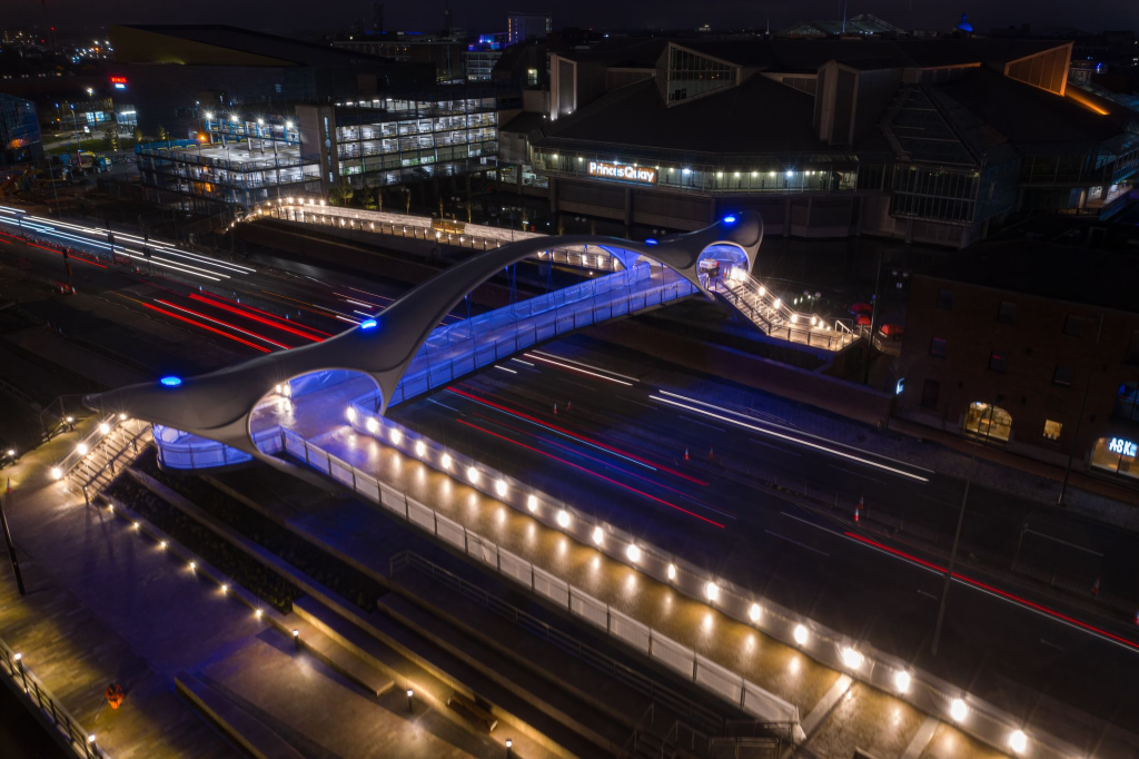 Aerial image of lit-up bridge at nighttime. The bridge is lit with blue lights and spans a lit up dual carriageway