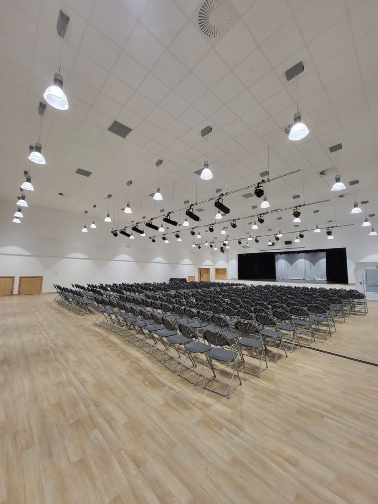 A large school hall with rows of seating in place facing a stage. There are stage lighting bars on the ceiling.