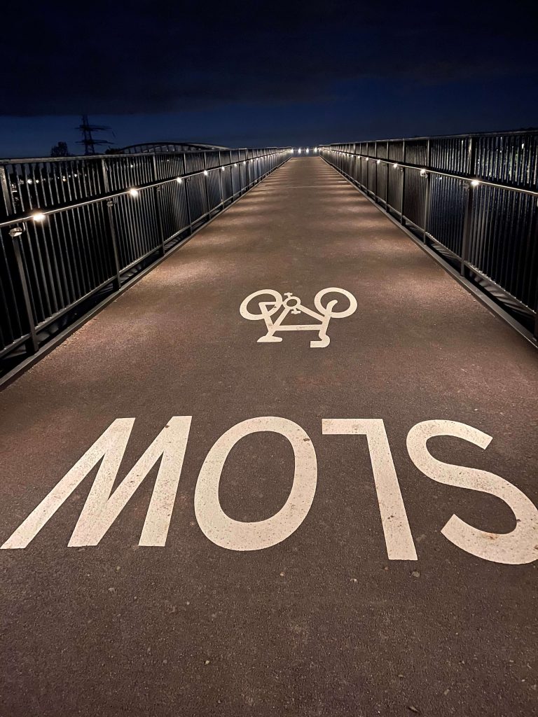 view of a bridge walkway with lighting on the handrails at nighttime. There is an inverted SLOW sign and bike painted in white on the pathway.