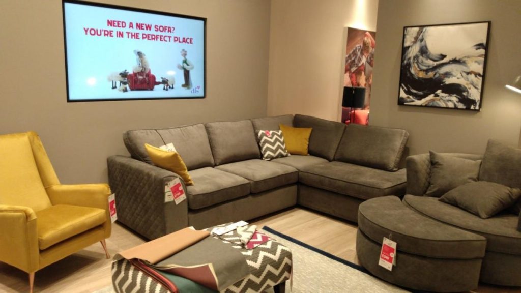 Living room setting in a furniture shop with a grey corner sofa, yellow armchair and grey ottoman. there is a picture on one wall and tv mounted on the other wall.
