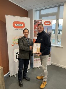 Two men standing side by side shaking hands. One man is holding a certificate.