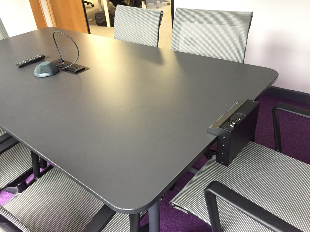 A table with five chairs in an office setting with AV equipment embedded into the centre of the table.