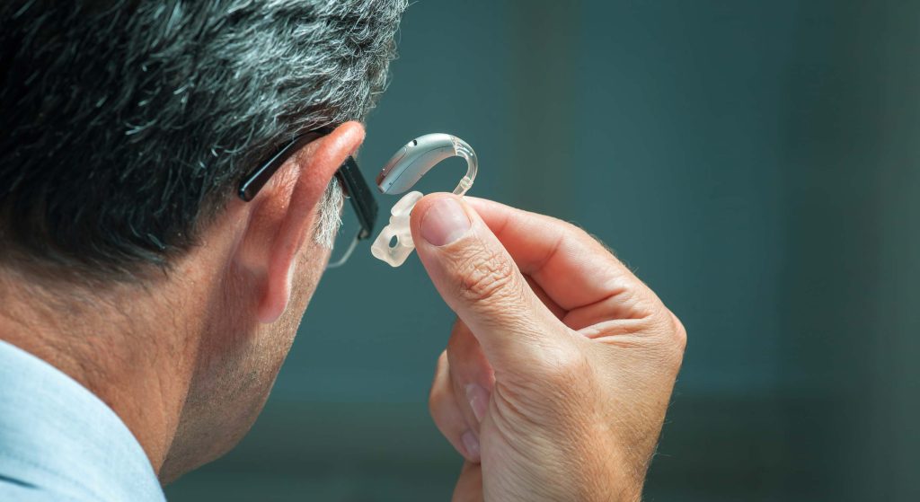 Close up of the back of a man's head. He is holding a hearing aid up to his right ear.