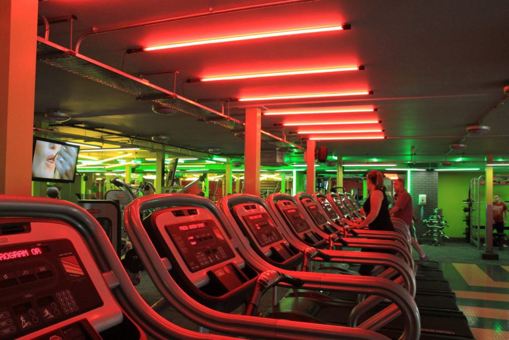 Interior shot of a gym focusing on a row of treadmills. The lighting is dim but there are vibrant red lights above the treadmills