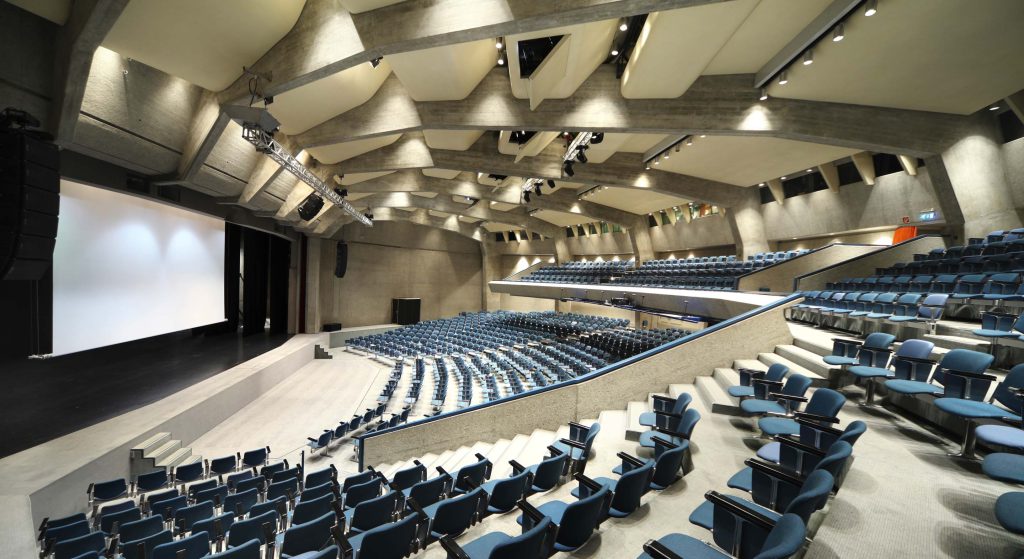 An empty lecture theatre, with blue seats arranged in a semi-circle facing the projection screen