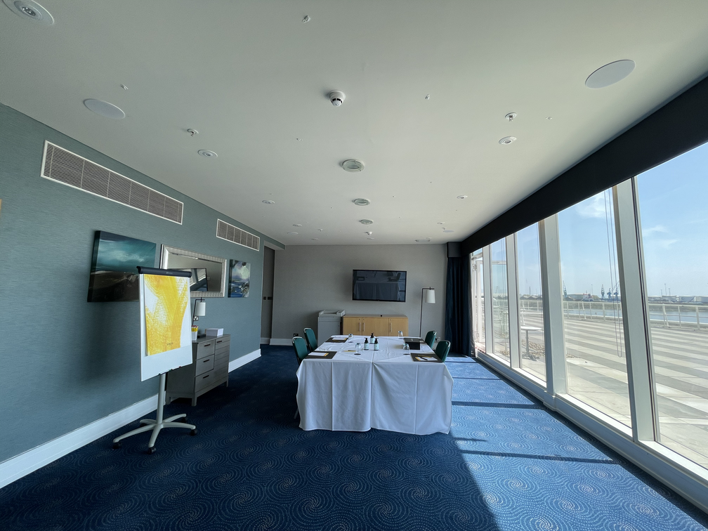 A sunny meeting room with a blue carpet, windows lining the right wall, and a table in the centre of the room