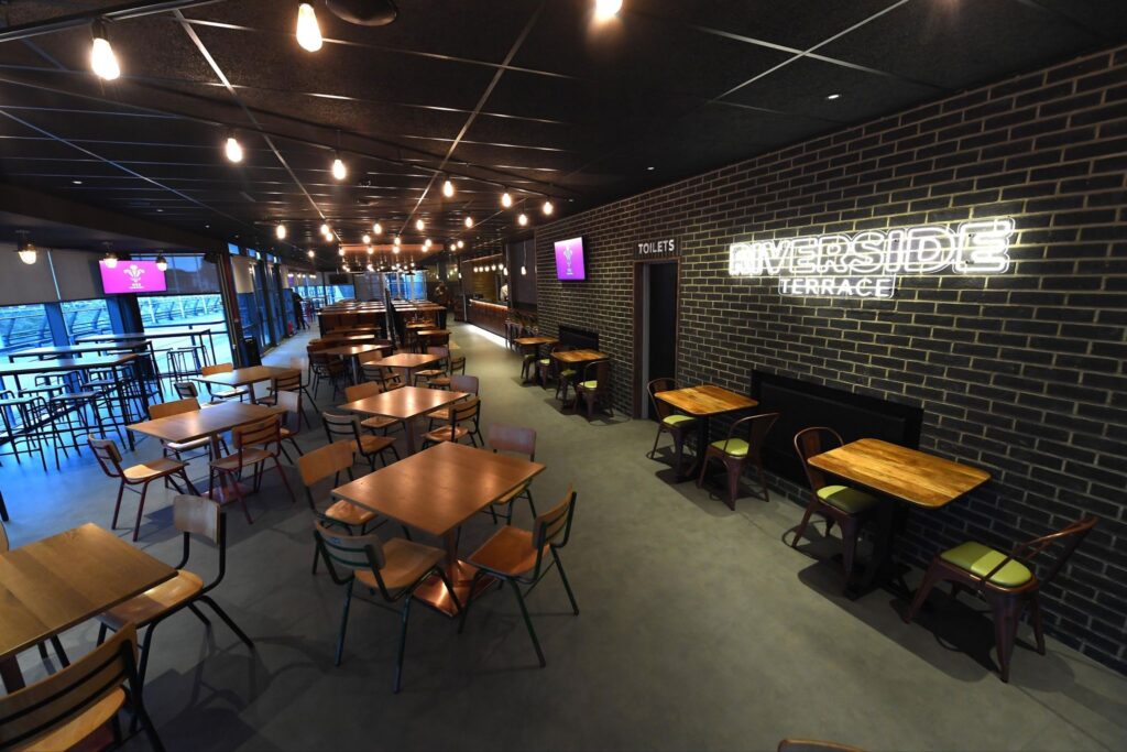 Interior of a bar with wooden tables of four in the foreground stretching out to the rear of the bar. There is a sign on the wall that says Riverside Terrace, lit up in neon lights.