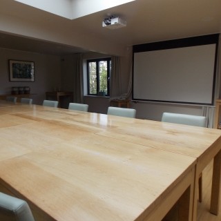 Image of boardroom setting in a hotel with a projector screen on the wall, a table in the centre of the image and five chairs either side of the table.