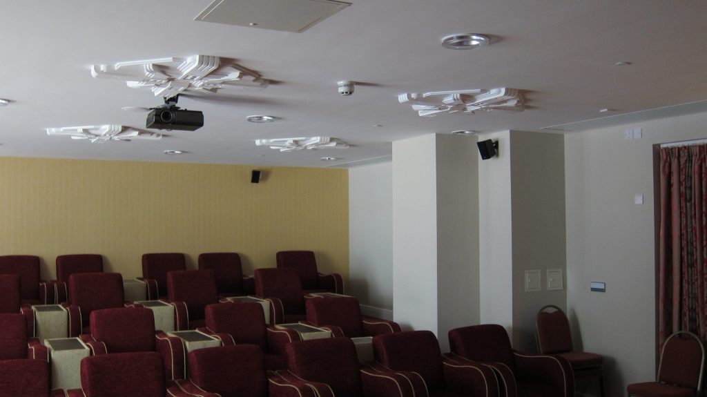 cinema room with dark red seats, a yellow wall in the background and a projector mounted to the ceiling.
