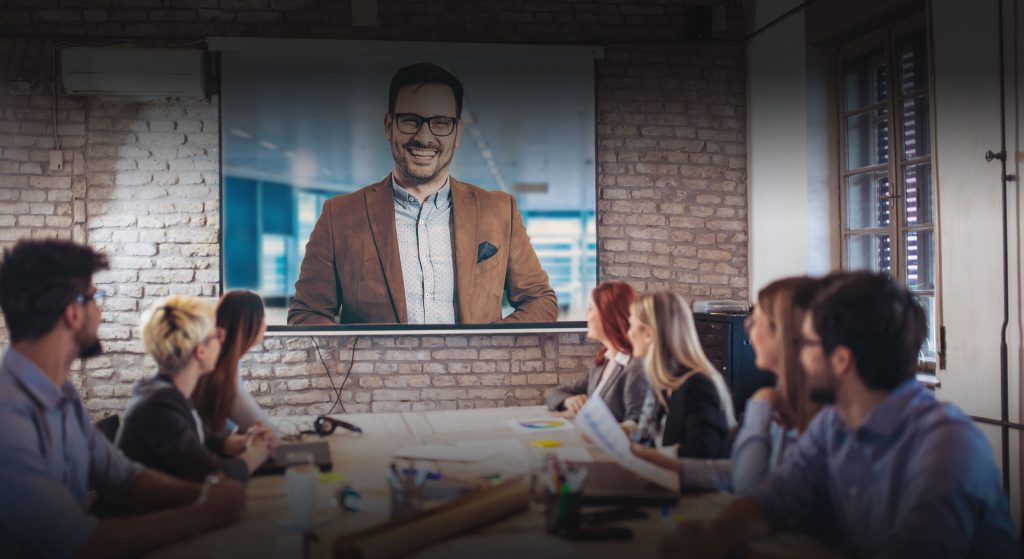 A team of people gathered around a meeting table, all turned to see a man speaking to them via a large display screen